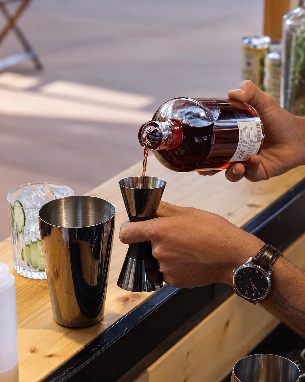 Un geste précis de dosage au jigger, liquide rouge foncé versé depuis une carafe, sur un comptoir en bois.