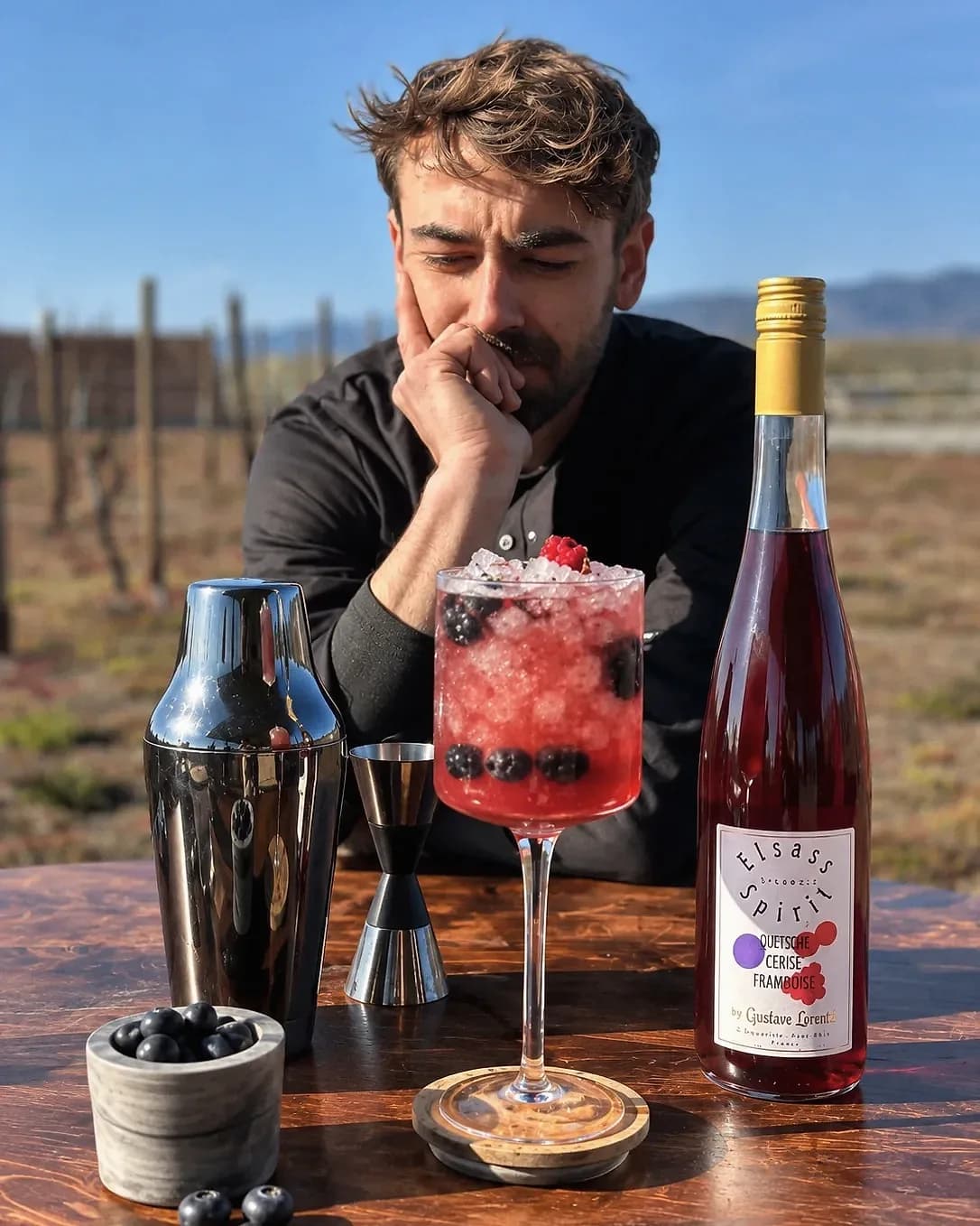 Lukas Guyot, le menton dans la main, devant une création aux fruits rouges, un shaker, un jigger et une bouteille Elsass Spirit, sur une table au milieu des vignes.
