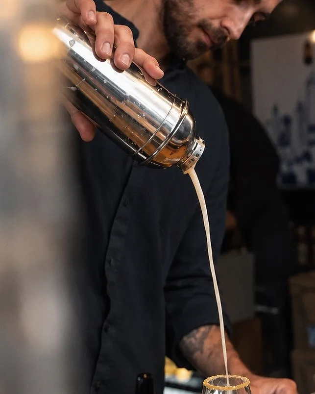 Lukas Guyot verse une préparation crémeuse d'un shaker dans un verre au bord sucré, derrière un comptoir de bar.