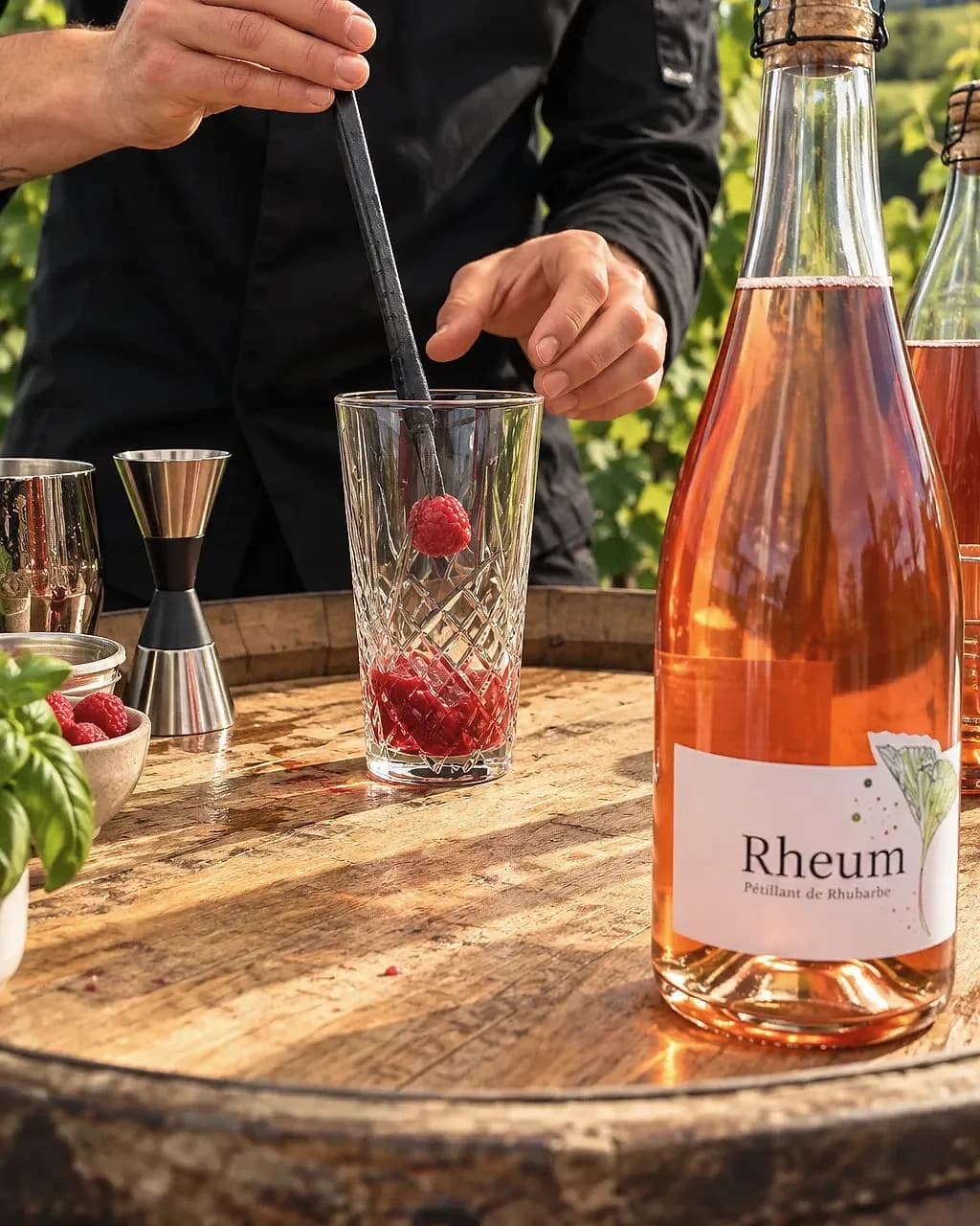 Des mains garnissent un verre de framboises fraîches, bouteille Rheum Pétillant de Rhubarbe du domaine, au milieu des vignes.