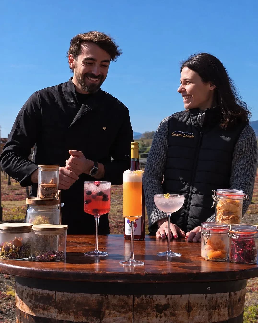 Un mixologue et une vigneronne de la maison Gustave Lorentz sourient devant trois créations et des bocaux d'épices et de fruits secs, sur un tonneau au milieu des vignes.
