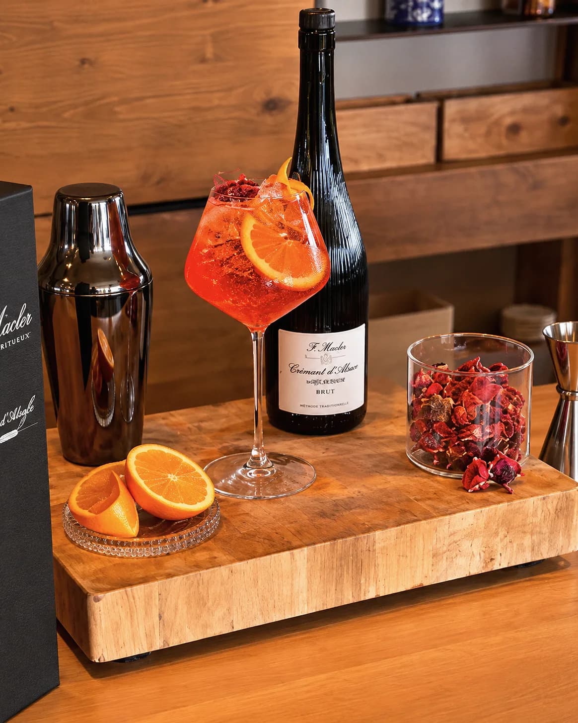 Une création cocktail rouge vif garnie d'un zeste d'agrume, devant une bouteille de Crémant d'Alsace F. Macler et un shaker.