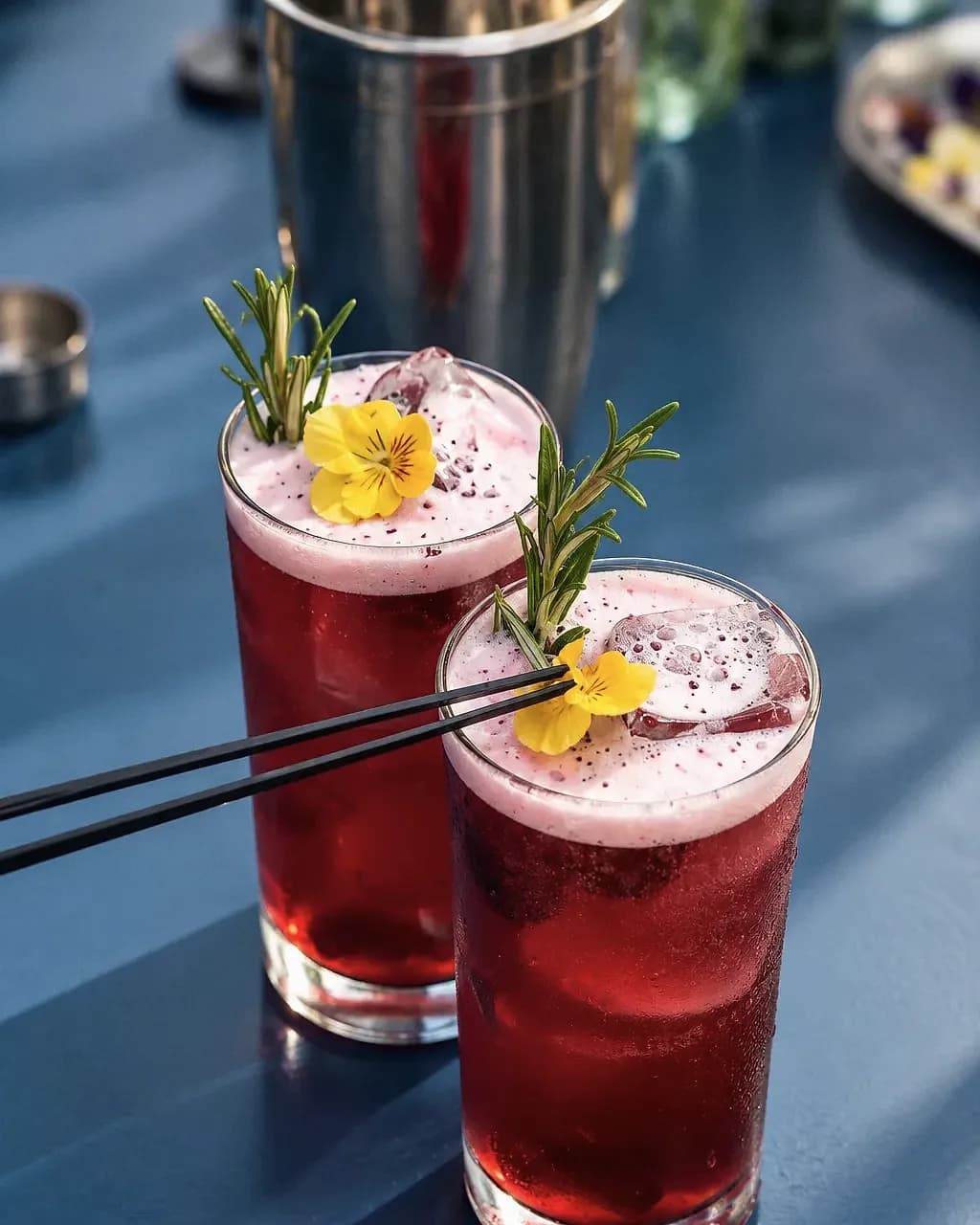 Un cocktail rouge garni d'une fleur et de romarin, dans un verre rempli de glace pilée.