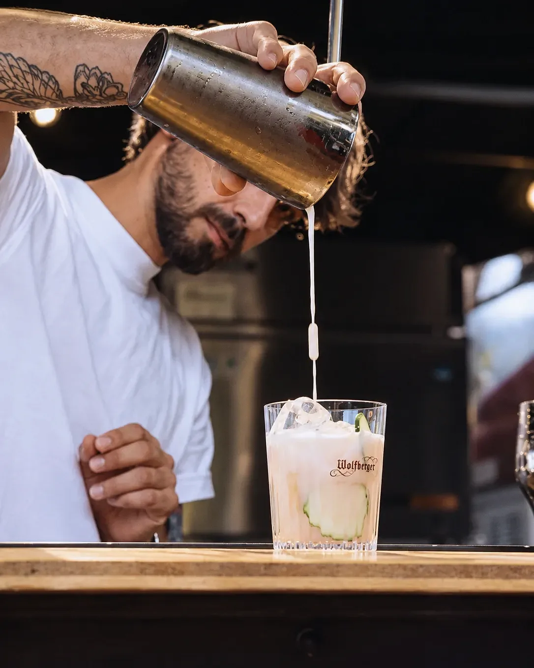 Un geste de service au shaker au-dessus d'un verre gravé Wolfberger, glaçons et rondelles de concombre.